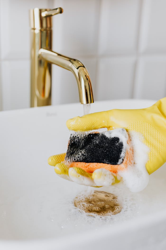home-hero Close-up of a yellow-gloved hand cleaning with a foamy sponge under a gold faucet in a sink.