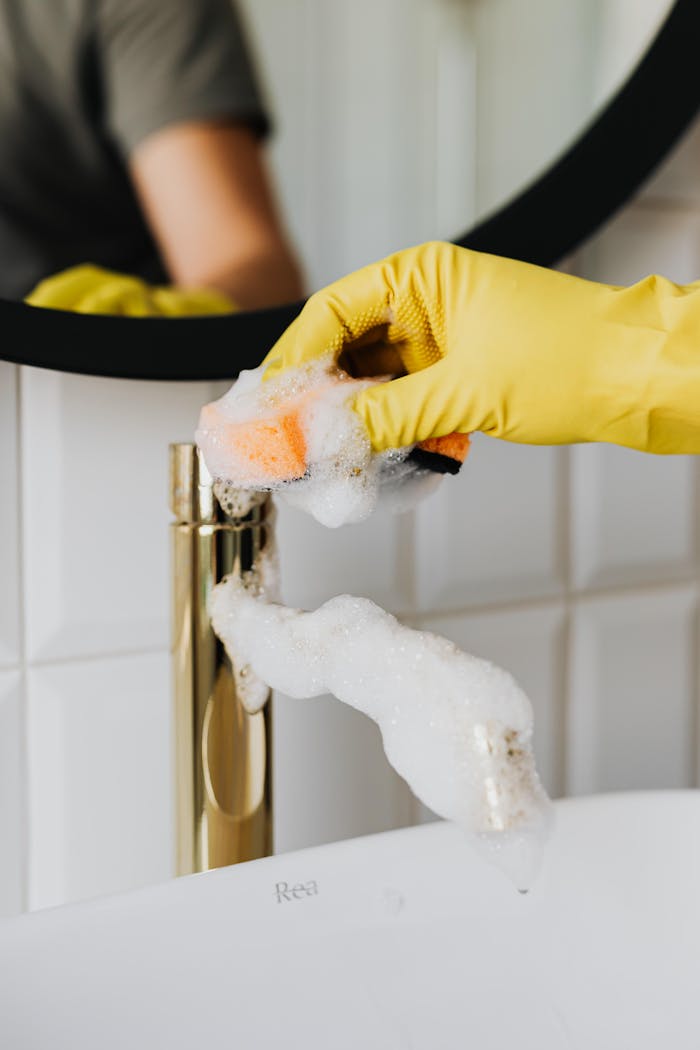 about-us Close-up of a hand in a yellow glove cleaning a bathroom faucet with foam.